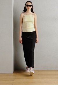 Lys gul ribbet singlet, svart gulvlengde skjørt, hvite klumpete sandaler, modell iført solbriller, med langt hår foran en ensfarget bakgrunn.