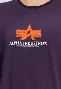 Fialové bavlněné tričko s oranžovým texturovaným logem s křídly a nápisem "Alpha Industries" potisknutým níže, s čistým a neformálním designem.