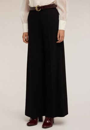 Mujer vestida con pantalones de pierna ancha de talle alto en color negro, blusa blanca transparente, cinturón marrón con hebilla dorada y zapatos de tacón rojos oscuros.