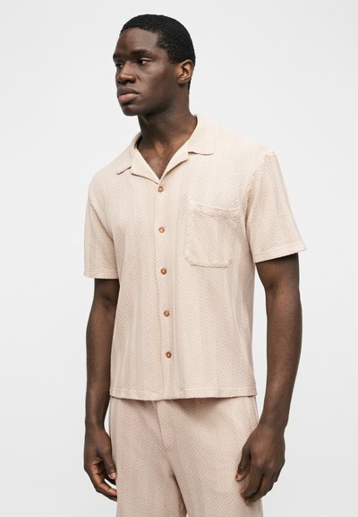 Homme portant une chemise beige à manches courtes avec poche poitrine et un pantalon assorti, debout devant un fond uni.