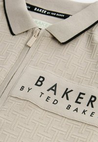 Polo beige con zip in tessuto testurizzato caratterizzato da un motivo intrecciato. Collo e dettagli neri. "BAKER by TED BAKER" stampato su un'etichetta.