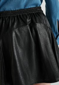 Zwarte faux leren rok met een elastische tailleband. Het materiaal heeft een gestructureerde afwerking en is geplisseerd. Een blauwe denimbloes is gedeeltelijk zichtbaar.
