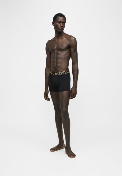 Homme musclé debout pieds nus, portant un boxer noir avec une ceinture à motif clé grec dorée sur fond blanc uni.