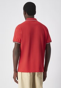 Rotes Poloshirt mit kurzen Ärmeln, kontrastierendem dünnem weißen Kragen und Akzenten an den Ärmeln. Der Stoff wirkt glatt mit einer Standardpassform und seitlichen Belüftungen.