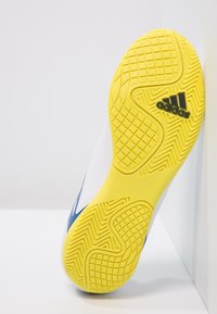 Vit sportssko med en texturerad gul gummisula. Blå detaljer på sidan, med ett hexagonalt mönster på det övre materialet.