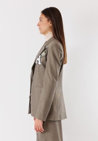 Blazer grigio su misura con revers a farfallino, caratterizzato da un'evidente toppa ricamata e una texture liscia, indossato sopra un leggero dolcevita.