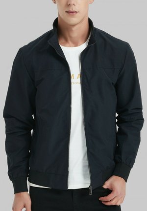 Veste zippée noire en tissu lisse, dotée d'un col montant, de poignets élastiques et d'un design minimaliste. Portée par-dessus une chemise blanche.