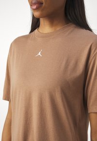 Camiseta de algodón marrón con mangas cortas, escote redondo y un logo blanco de Air Jordan en la parte superior del pecho. Textura suave, corte casual.