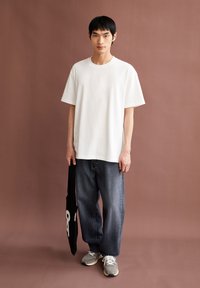 T-shirt blanche à manches courtes avec un col rond, associée à un jean large bleu foncé et des baskets grises. Sac fourre-tout noir avec un graphique blanc.