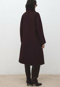 Bordeaux wollen jas met een hoge kraag, lange mouwen en een rechte snit. Draagt bij glossy bruin laarzen tot op de knie.