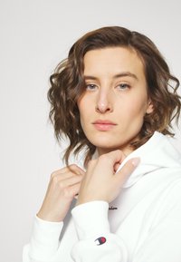 Felpa bianca con cappuccio e cordino, con logo ricamato sul petto. Il tessuto appare morbido. Il modello ha capelli ricci fino alle spalle.