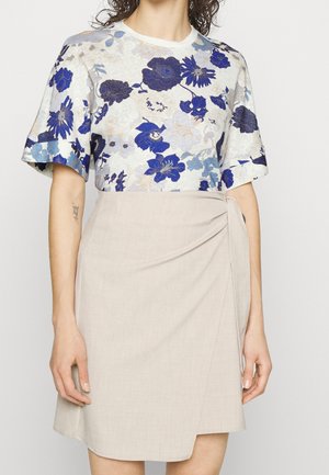 Kobieta ubrana w białą bluzkę z niebieskimi kwiatowymi wzorami i beżową spódnicę kopertową, stojąca na tle jasnego, jednolitego tła.