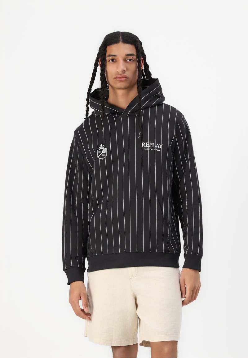 Svartstripete hettegenser med frontlomme, med hvite striper og logo. Kombinert med lysebeige shorts. Jevn tekstur.