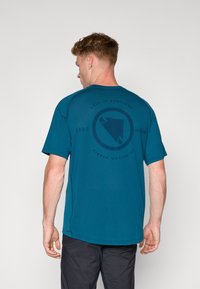 T-shirt turquoise avec un imprimé rond sombre au dos comportant le texte « Né en Écosse » et un élément de design. Manches courtes et texture douce.