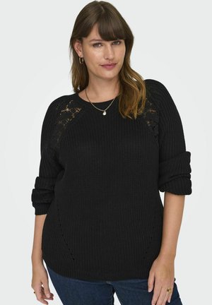 Sort strikket sweater med et struktureret mønster, rullede manchetter og sideslids. Sammen med blå jeans og en enkel guldhalskæde.