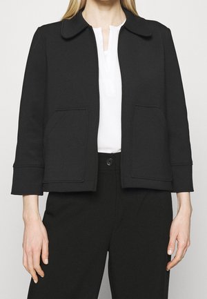 Zwarte cropped jas met een ronde kraag, twee voorzakken en gestructureerde mouwen. Draagt over een witte top en zwarte broek.