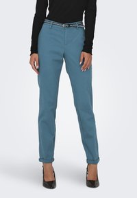 Zilas kokvilnas bikses ar sašaurinātu piegriezumu, sarullētām piedurknēm un kontrastējošu melnbaltu svītrainu jostu, kombinētas ar melniem papēžiem.