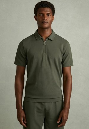 Man draagt een olijfgroene polo met korte mouwen en een ritssluiting aan de voorkant, bijpassende broek, staande tegen een effen grijze achtergrond.