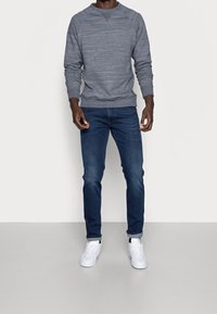 Sweat-shirt gris texturé avec un col rond, associé à un jean bleu foncé et des baskets blanches, avec des poignets retroussés et des éléments de design simples.