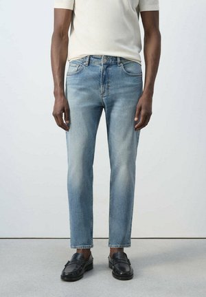 Hombre con jeans azul claro de corte recto, una camisa blanca de manga corta y mocasines de cuero negro, de pie frente a un fondo liso.