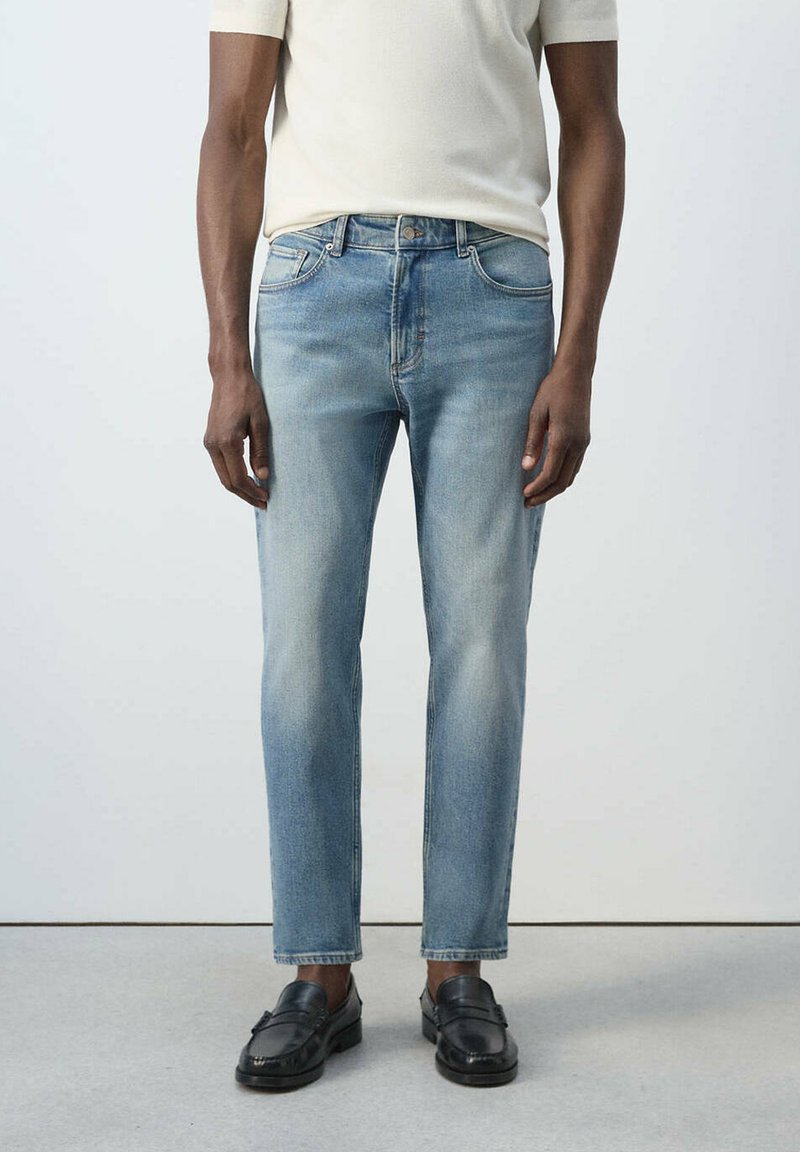 Homme portant un jean droit bleu clair, une chemise blanche à manches courtes et des mocassins en cuir noir, debout devant un fond uni.