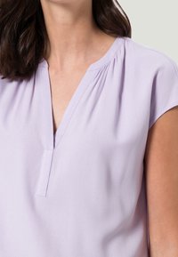 Blouse lavande avec un décolleté en V, des manches courtes, un tissu doux et des plis subtils le long du décolleté pour un détail supplémentaire.