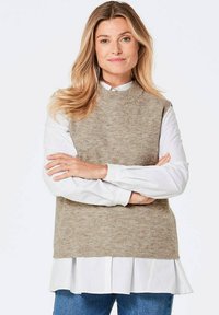 Pull en tricot sans manches marron clair porté par-dessus une chemise blanche à col, avec un ourlet froncé, associé à un jean bleu. Texture lisse et design à côtes.