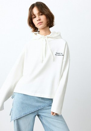 Jeune femme en sweat à capuche blanc avec le texte « Voyage de la mer, du sable et du soleil », portant une jupe en denim bleu clair asymétrique et un jean.