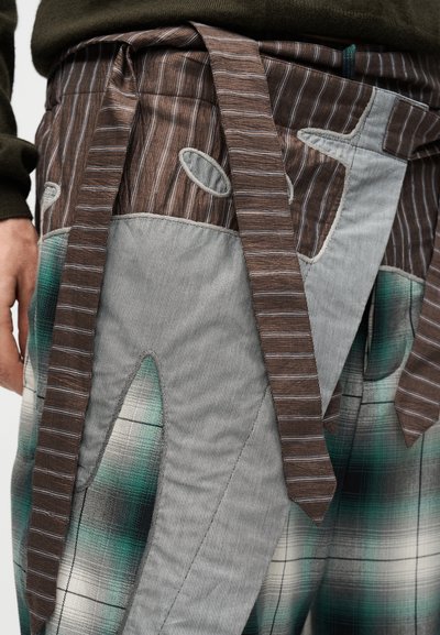 Κοντινό πλάνο ενός ατόμου που φορά πολύχρωμο παντελόνι patchwork με ριγέ καφέ ζώνη και πράσινο καρό μοτίβο, χέρι ακουμπισμένο στο πλάι.