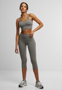 Grå treningscrop top med kryssede stropper, kombinert med høy midje grå tights. Modellen har på seg grå joggesko. Glatt, stretchy materiale.
