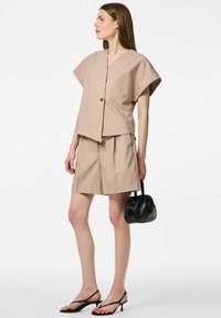 Top beige court à manches courtes avec un bouton sur le devant, associé à un short beige sur mesure. Le mannequin tient un petit sac à main noir.