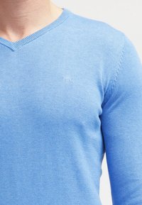 Pull en V bleu clair fabriqué en maille douce, présentant un discret logo et une coupe ajustée. Texture lisse sans motifs.