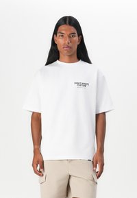Weißes Kurzarm-T-Shirt mit einem kleinen, schwarzen "DON'T WASTE CULTURE" Textlogo auf der linken Brust. Weicher Stoff, lockere Passform.