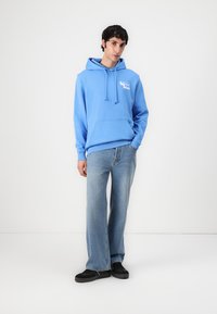 Lichtblauwe hoodie met een voorzak, gemaakt van katoen, met trekkoorden; gecombineerd met lichtblauwe brede jeans en zwarte schoenen.