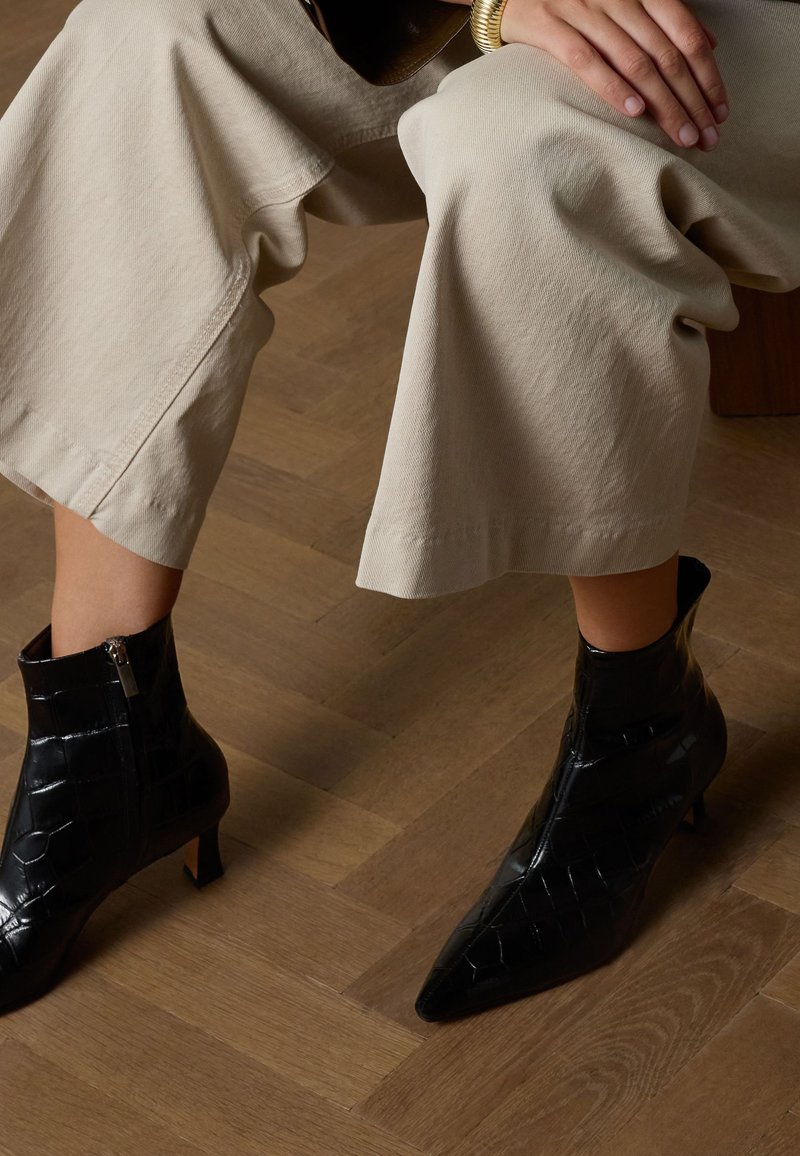 Black ankle boots with pointed toes, textured crocodile pattern, zipped side closure, and a small heel, paired with beige wide-leg pants.