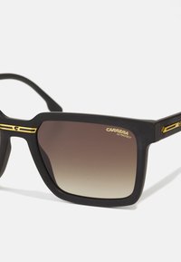 Occhiali da sole rettangolari neri con finitura opaca, lenti gradientate marroni e dettagli dorati sulle tempie. Presentano il marchio "CARRERA UV PROTECT".