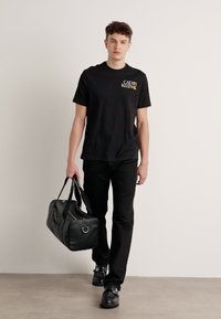 Sort bomulds T-shirt med gult logo, parret med sorte bukser og sko, mens han/hun holder en sort læder duffel taske med sølv hardware.