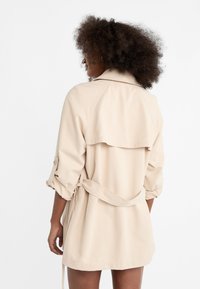 Trench-coat beige avec une ceinture à nouer, des manches retroussées et un dos à rabat superposé. Tissu lisse, coupe au genou, avec des détails de couture minimalistes.