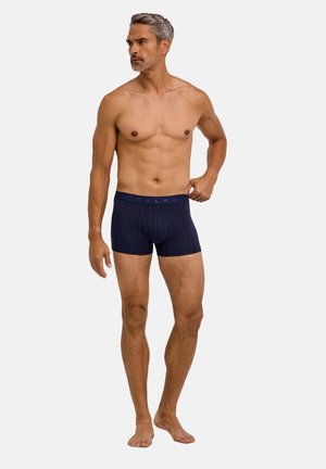 Hombre musculoso con cabello gris que lleva calzoncillos tipo bóxer azul oscuro con estampado, de pie descalzo sobre un fondo sencillo y claro.