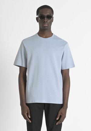 Lichtblauw katoenen T-shirt met korte mouwen en een ronde hals. Heeft een subtiel logo geborduurd op de borst. Zwarte broek en zonnebril.