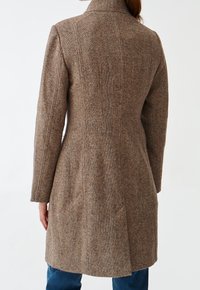 Abrigo de tweed marrón con forma entallada, cuello alto y mangas largas. Presenta una tela texturizada con sutiles motas de colores y costuras traseras.