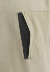 Tissu beige avec un tissage texturé, orné d'une poche zippée noire contrastante. La poche a une surface douce et lisse.