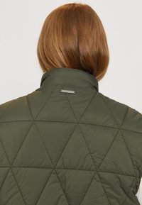 Olivgrüne gesteppte Jacke mit Rautenmuster, ausgestattet mit einem Stehkragen und einem kleinen metallischen Marken-Tag auf der Rückseite. Glatte Stofftextur.