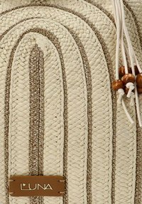 Bolsa de mimbre beige con patrones texturizados, acentuada por hilo dorado y cuentas de madera, con una etiqueta de logo marrón. Cierre de cordón en la parte superior.