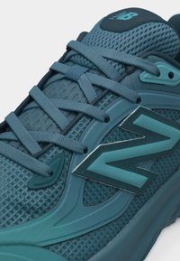 Κοντινό πλάνο αθλητικού παπουτσιού New Balance σε τιρκουάζ χρώμα, με υφή από πλεκτό δίχτυ, κορδόνια στο ίδιο χρώμα και εμφανές λογότυπο της μάρκας στο πλάι.