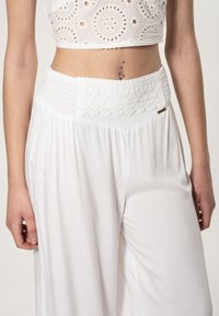 Witte wijde broek met een gestructureerde, smocktailband, voorzien van kanten details en een kleine metalen accent op de heup.