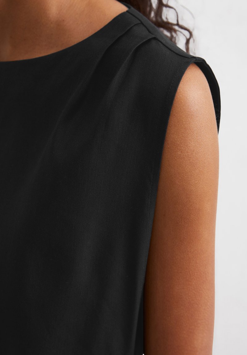 Schwarzes ärmelloses Kleidungsstück aus einem glatten, weichen Stoff mit rundem Halsausschnitt und dezenten Schulterfalten. Die Textur wirkt leicht und fließend.