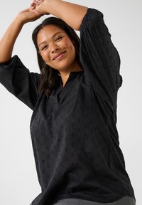 Chemise noire à coupe ample, avec col mandarin, manches trois-quarts et un motif texturé de petites formes sur l'ensemble.
