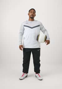 Grijze sportshirt met lange mouwen en een donker chevronpatroon, zwarte sportbroek en witte voetbalschoenen met rode veters. Houdt een witte voetbal vast.