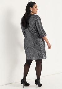 Kort paljettklänning i silver, med trekvartslånga ärmar, en åtsittande silhuett och en glänsande textur. Matchas med svarta strumpbyxor och klackar.